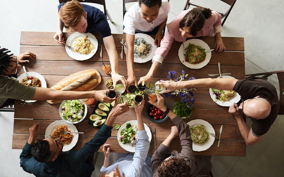 Middagsbjudning uppifrån med åtta vänner som skålar med glasen ovanför bord med mat och grönsaker.
