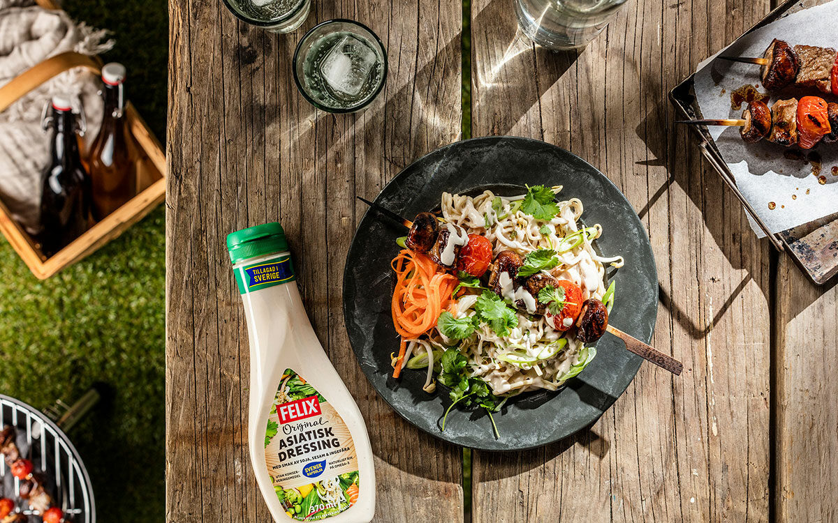 En tallrik med nudlar och grillspett på ett somrigt träbord, en flaska asiatisk dressing bredvid.