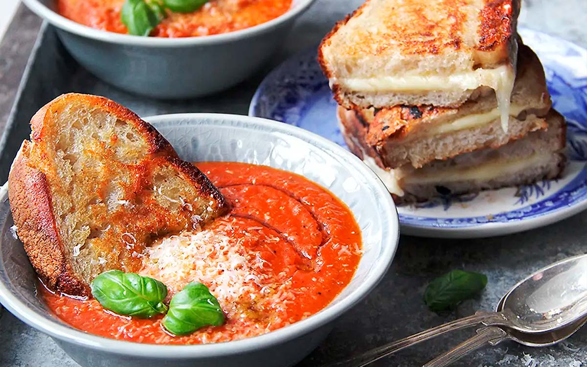 Skål med tomatsoppa toppad med basilika och pamesanost med grilled cheese mackor.