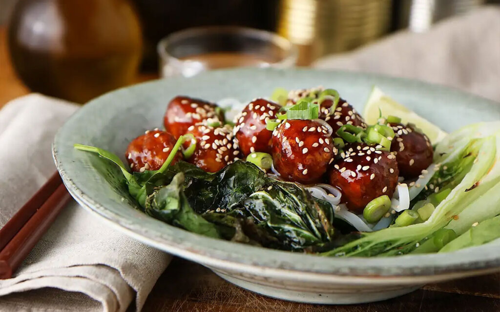 Tallrik med vegetariska delikatessköttbullar i teruyakisås på en bädd av risnudlar och pak choi toppad med skivad salladslök och sesamfrön.