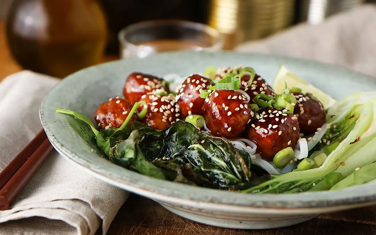Tallrik med vegetariska delikatessköttbullar i teruyakisås på en bädd av risnudlar och pak choi toppad med skivad salladslök och sesamfrön.